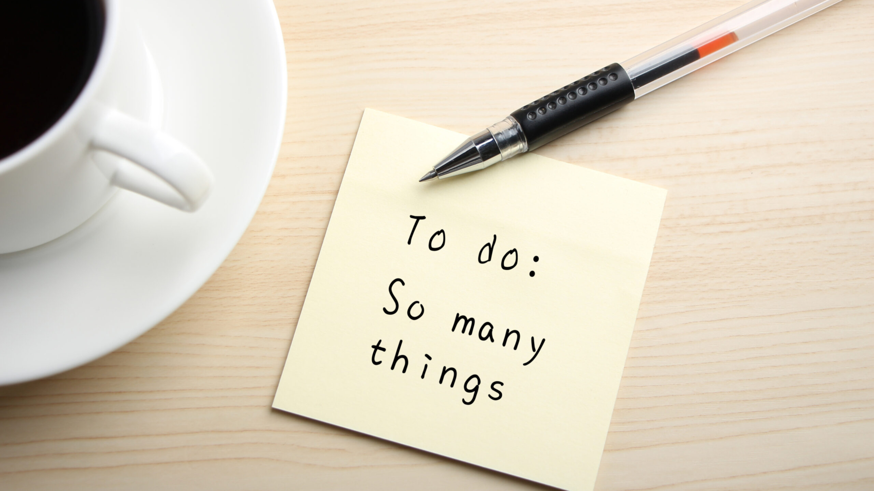 A yellow sticky note is written something on the desk with a cup of coffee and a ball pen aside.